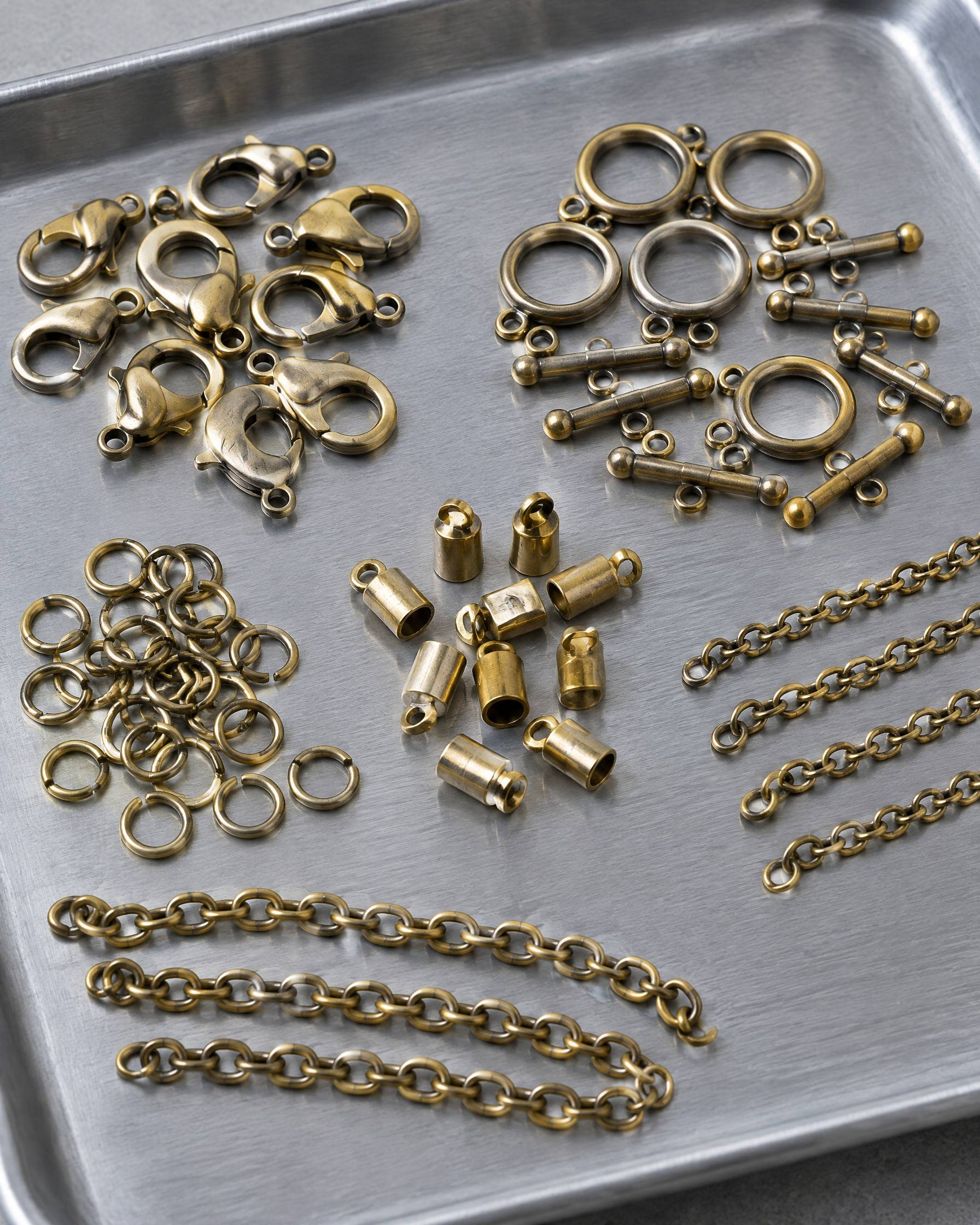 Raw brass jewelry components sorted on an aluminium tray: lobster clasps, toggle clasps, jump rings, end caps and chain.