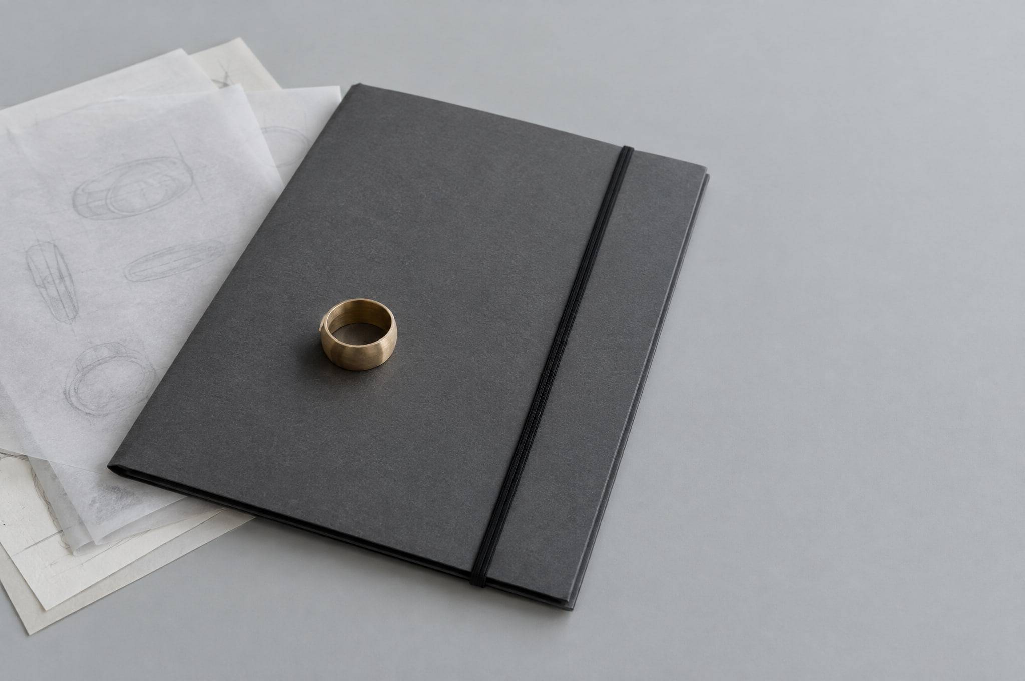 A closed grey portfolio held by an elastic band, design sketches turned face down under tracing paper, and a raw brass ring prototype.