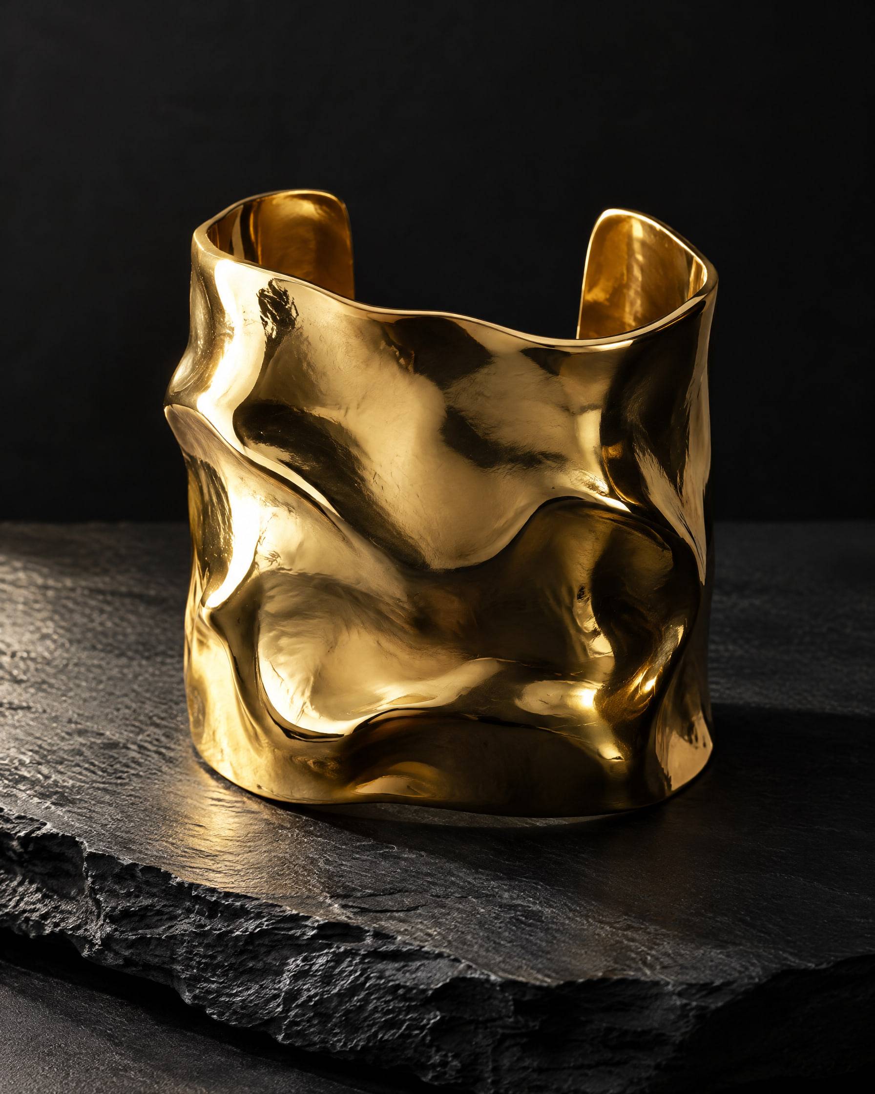 A wide sculptural gold-plated brass cuff with a molten, folded surface, standing on dark slate.
