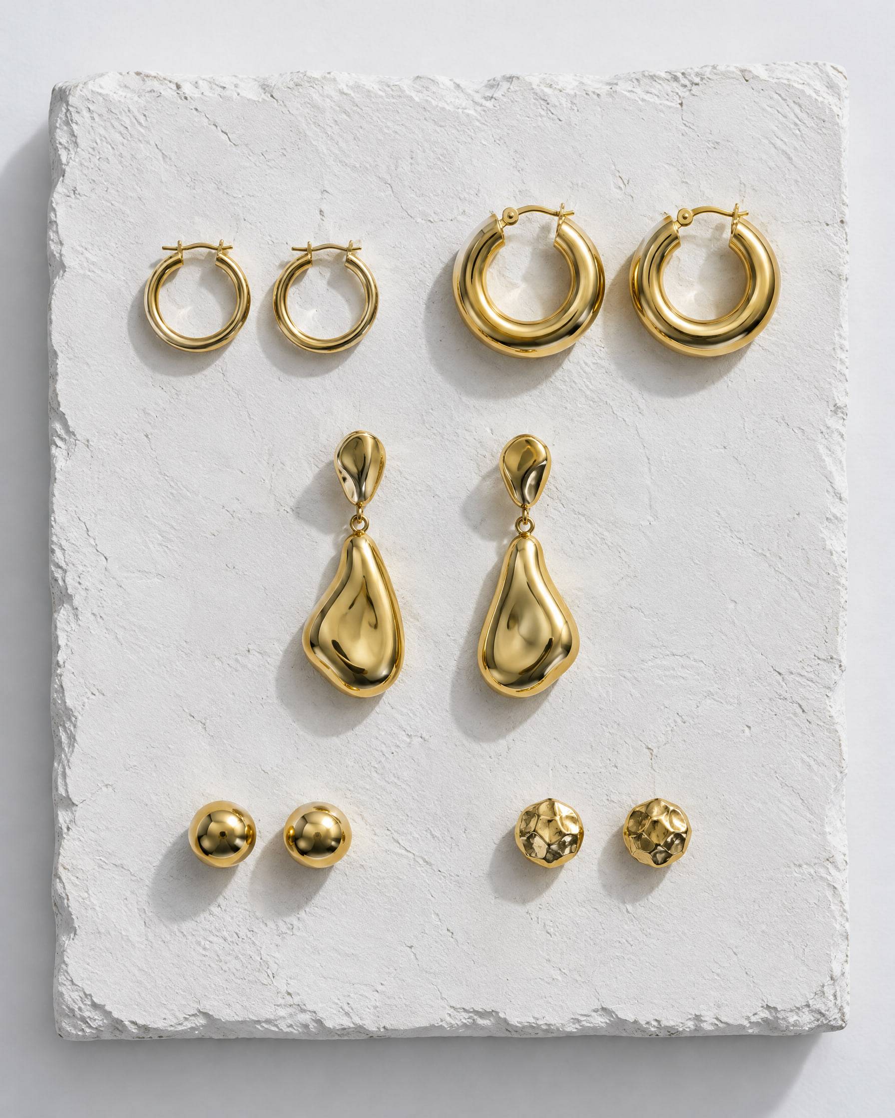 Five pairs of gold-plated brass earrings on a white plaster slab: small hoops, chunky hoops, organic drops and two pairs of studs.