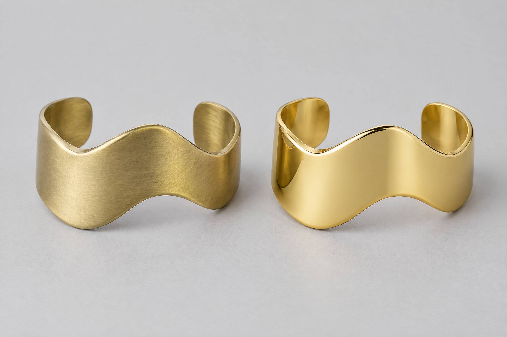The same wave-shaped cuff twice: on the left in raw satin brass, on the right gold-plated with a polished finish.
