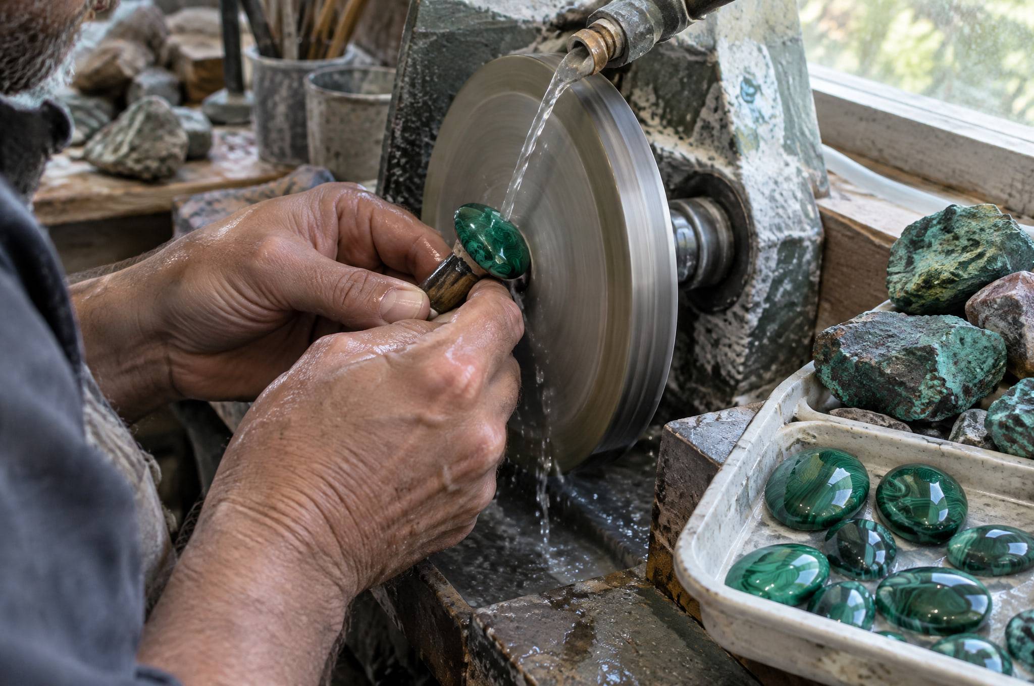 A lapidary shaping a malachite cabochon on a wet grinding wheel, with rough stones and finished cabochons beside the wheel.
