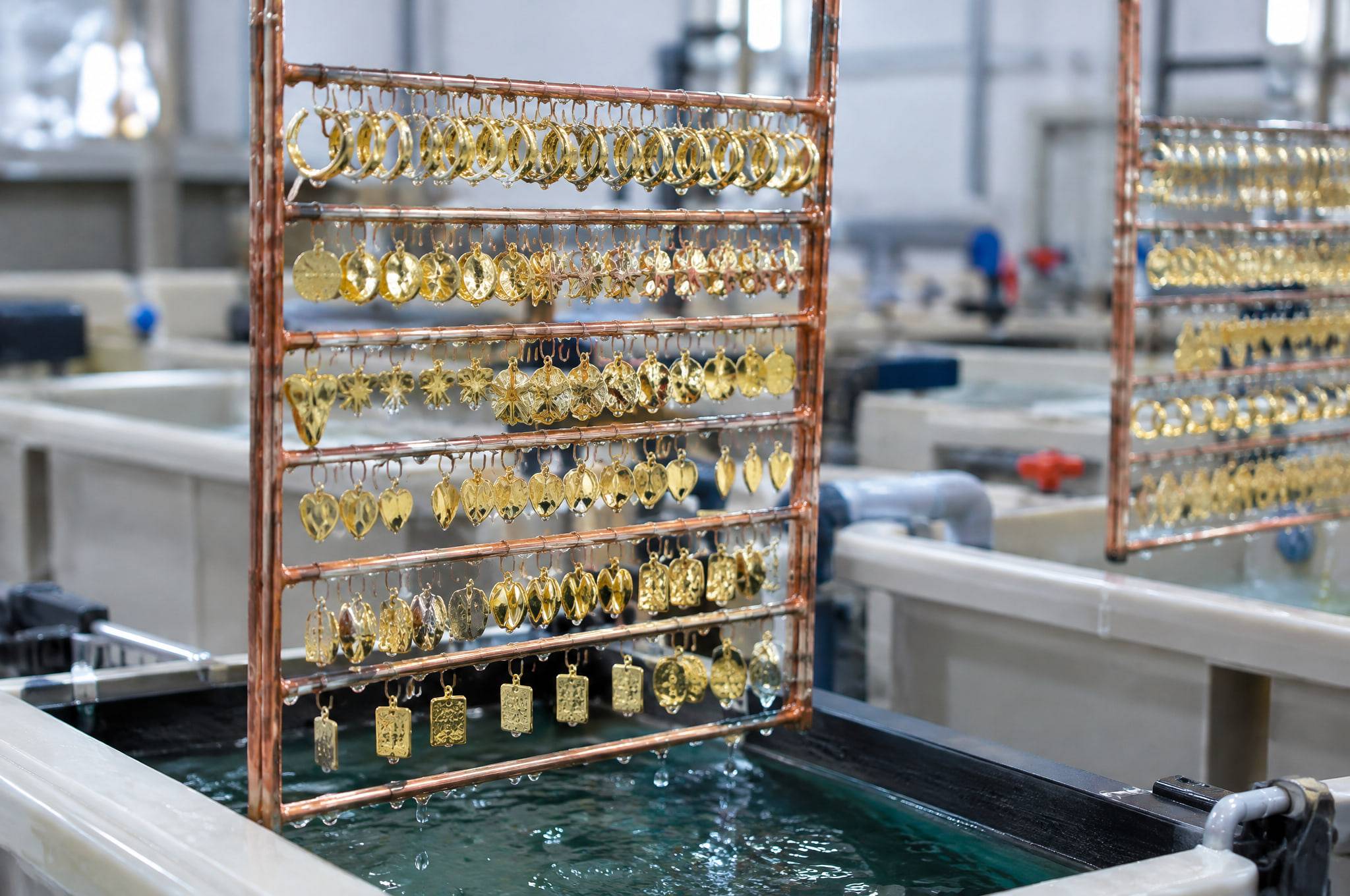 Freshly gold-plated earrings and pendants hanging in rows on copper plating racks above a plating tank.