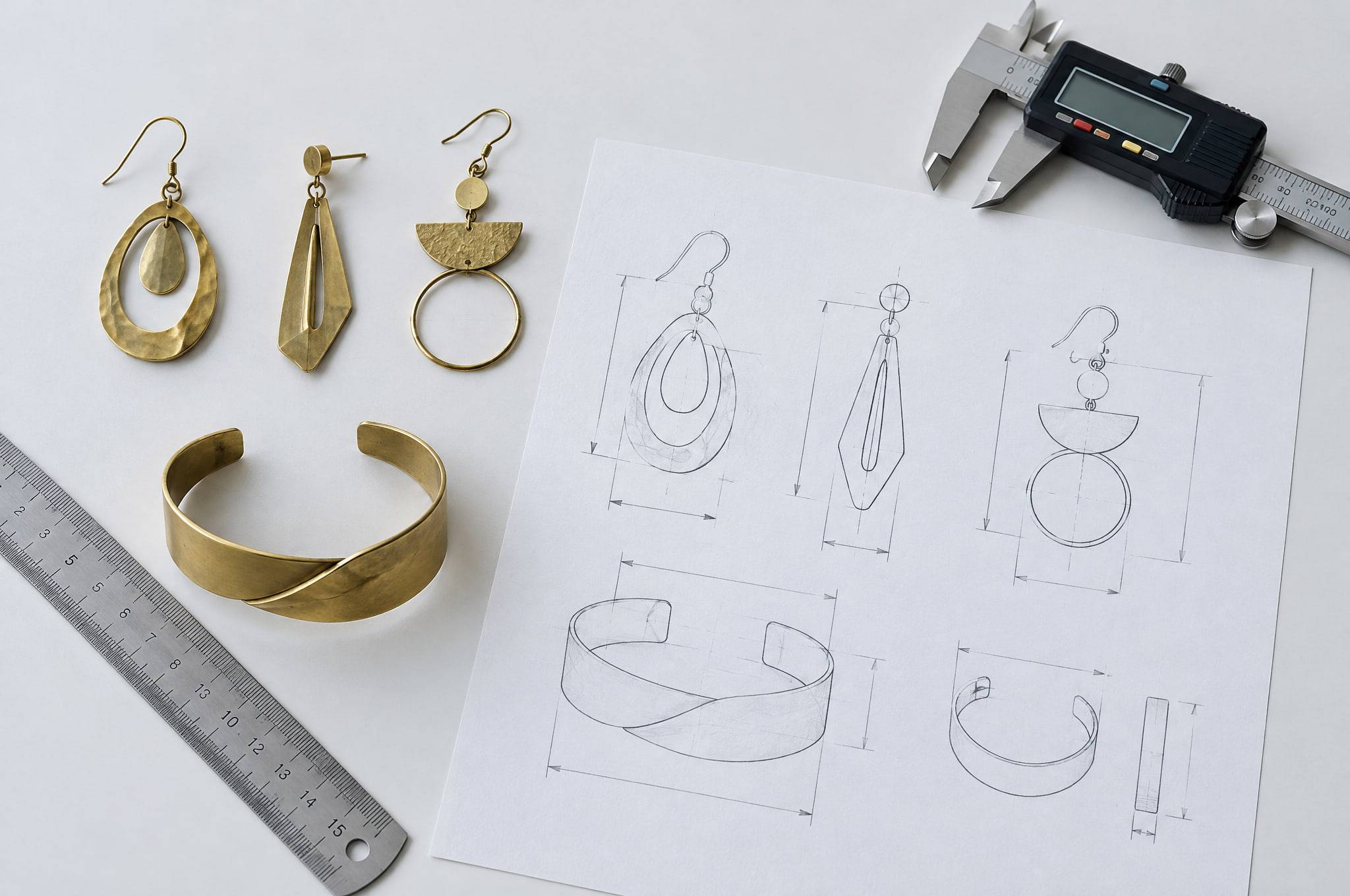 Raw brass prototype earrings and a cuff beside the pencil technical drawing they were made from, with a ruler and a digital caliper.