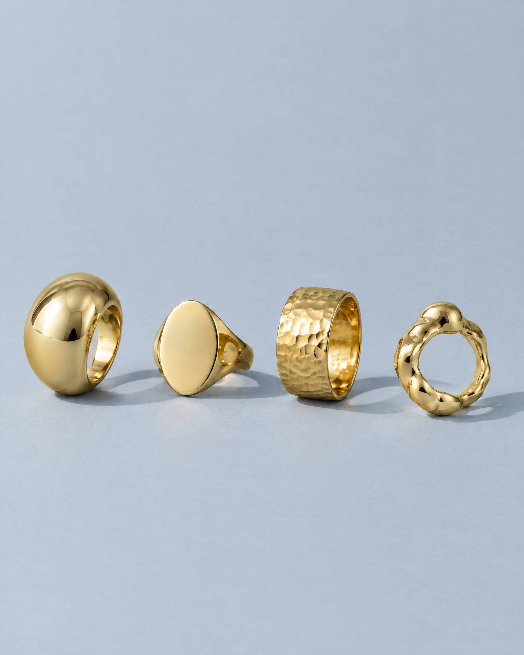 Four gold-plated brass rings on pale blue paper: a dome ring, an oval signet, a hammered band and an organic ring.