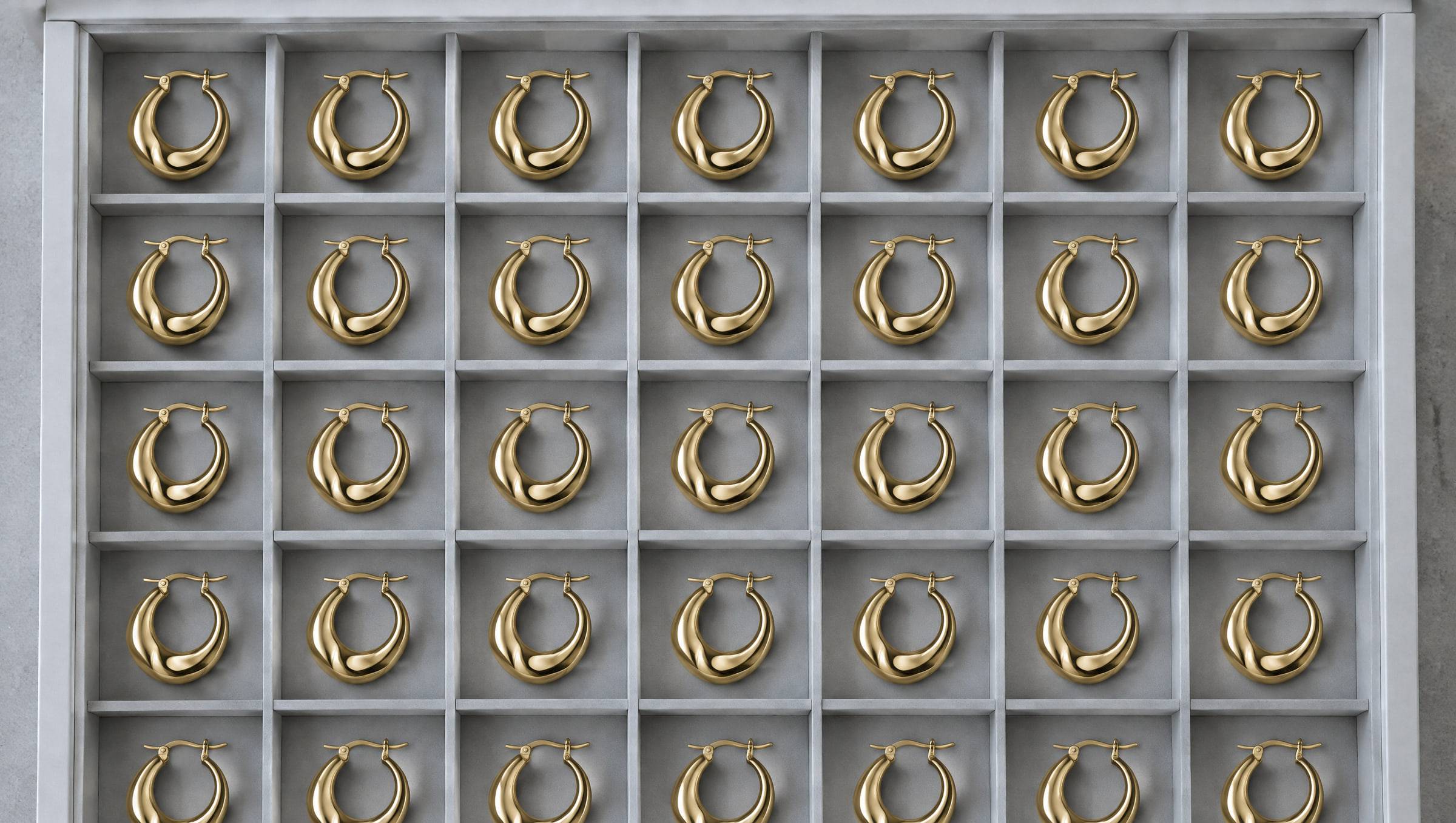A grey compartment drawer holding about forty identical gold-plated sculptural hoop earrings — one design, one small production run.