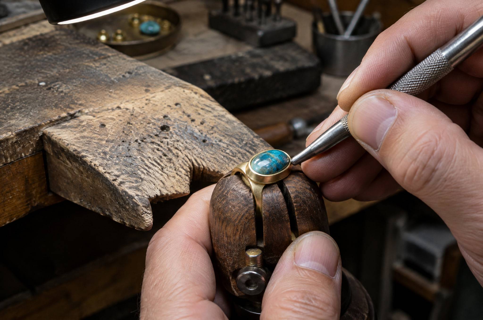 A jeweler's hands setting an oval blue-green cabochon into a gold-plated ring held in a ring clamp.