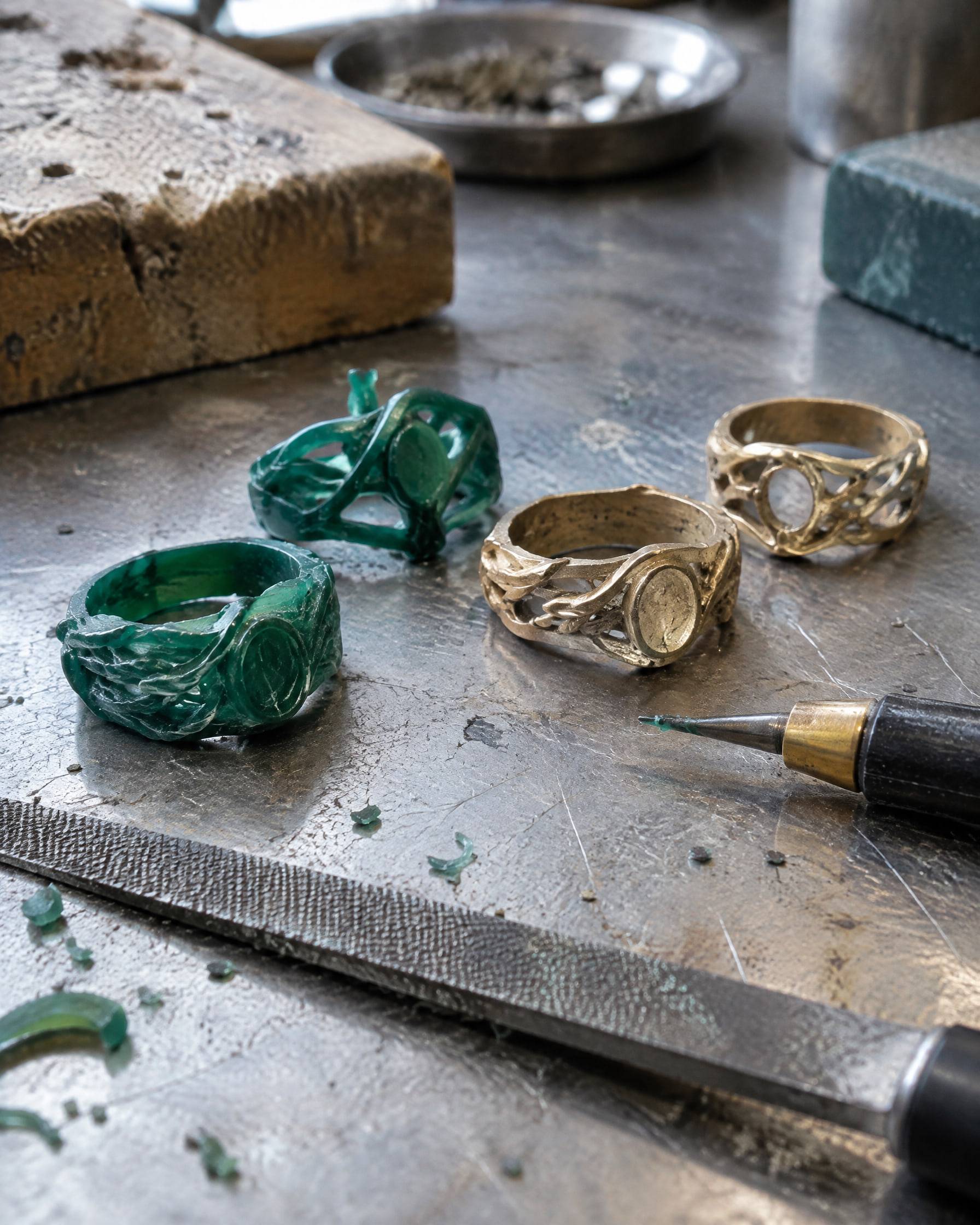 Carved green wax ring models beside two freshly cast raw brass ring prototypes on a steel workbench.