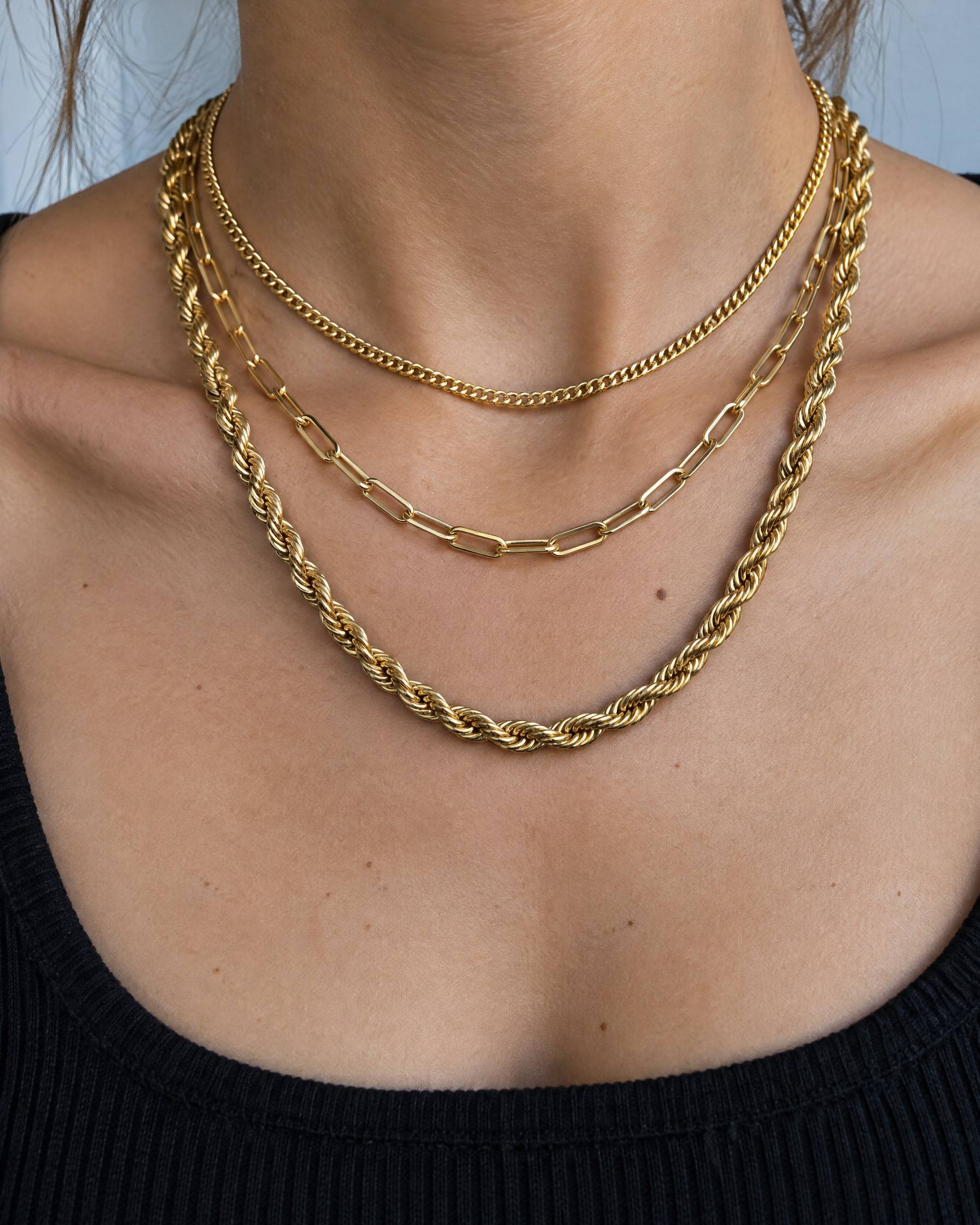A neck wearing three layered gold-plated brass chains: a fine curb chain, a paperclip chain and a rope chain.