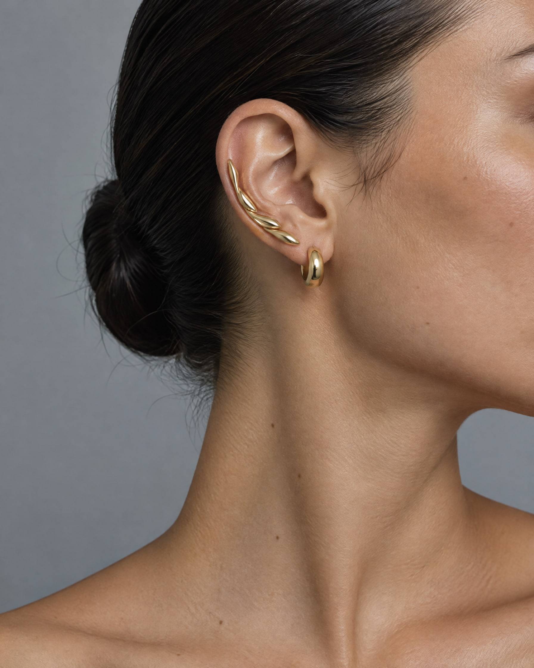 Profile of a woman wearing a sculptural gold-plated ear climber and a small chunky huggie hoop.