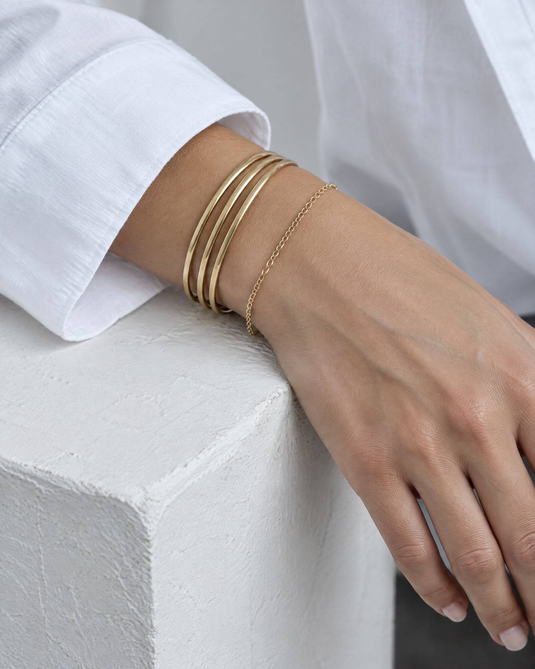 A wrist wearing three thin gold-plated bangles and a fine chain bracelet, resting on a white plaster block.